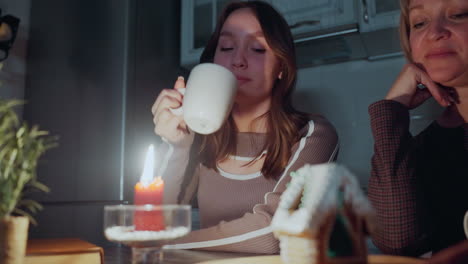 una cálida luz de vela brilla junto a una casa de pan de jengibre intrincadamente decorada en una bandeja de madera, una mujer bebe de una taza mientras otra mira pensando, en un entorno débilmente iluminado