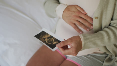 hands of caucasian pregnant woman sitting on bed, touching belly and looking on ustrasound photo