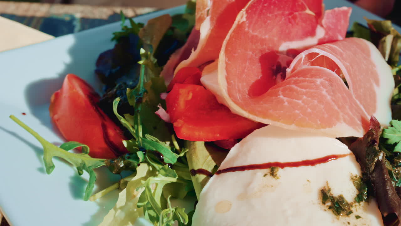 Close up of a gourmet salad with fresh mozzarella, cured meat slices, tomatoes, olives and mixed greens on a white plate in bright daylight