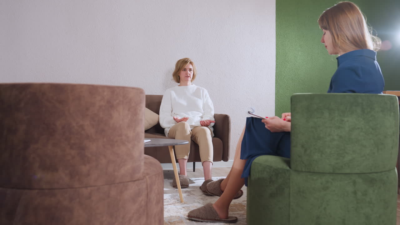 Zoomed view of guest speaking to professional as notes are taken by pen on pad, cozy therapy setting with green chair, brown sofa, textured walls, table with clock and pen pack, soft slippers visible