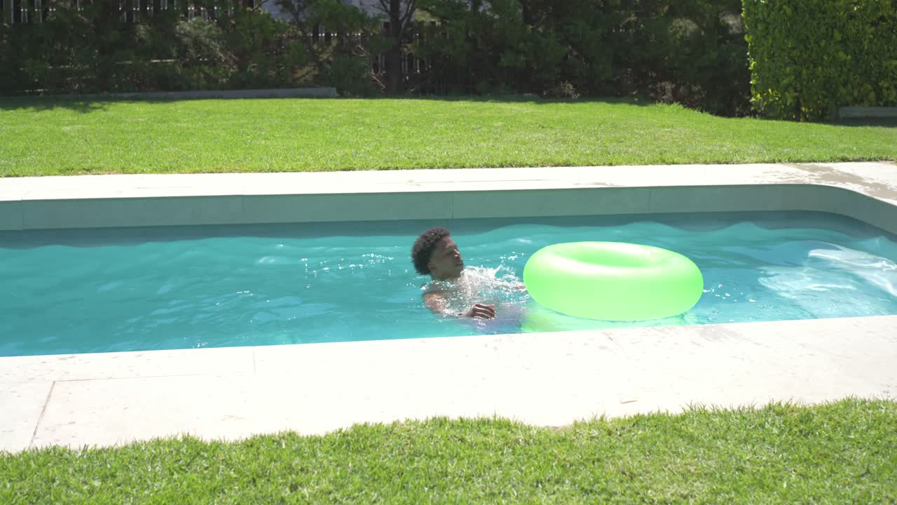 Adult man running, jumping from lawn into backyard pool, bright trunks, using neon ring cooling off