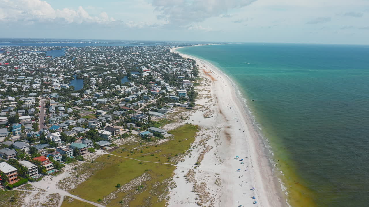 Rows of seaside homes and buildings stretch along a pristine sandy beach where turquoise waters meet the horizon, depicting a vibrant coastal lifestyle by the sea