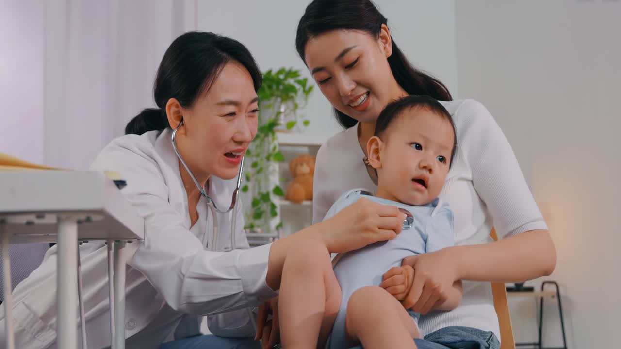 Female doctor examining cute boy with stethoscope. Baby happily smiling and pointing on doctor. Mother holding her kid. Pediatrician visit his patient at home
