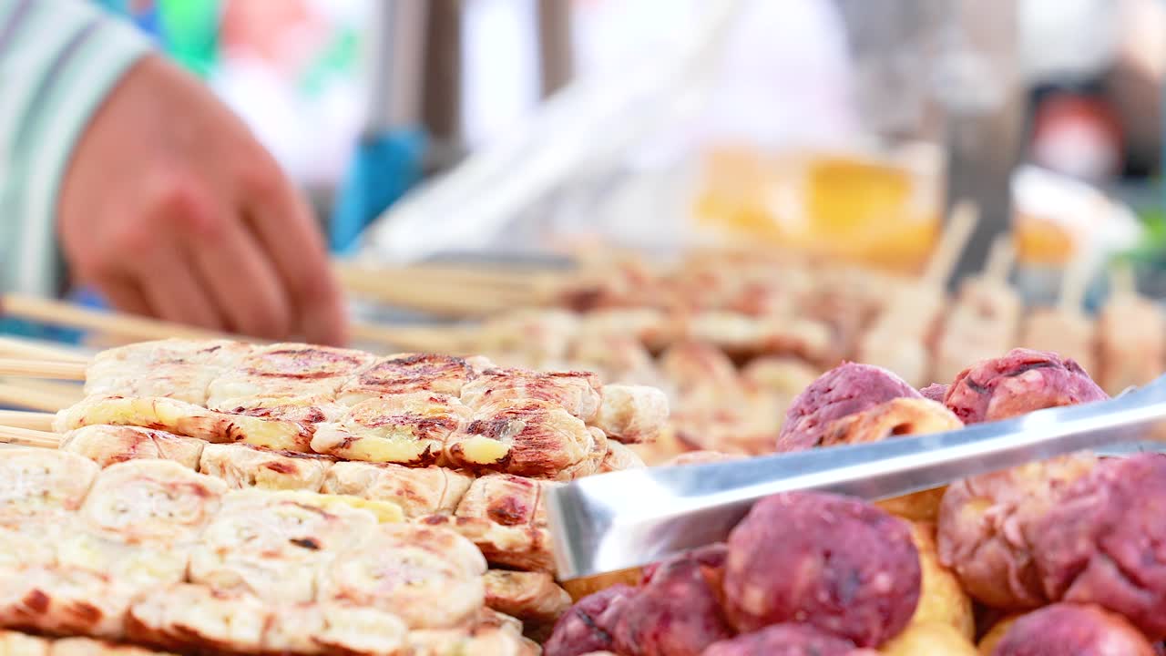A vendor grills banana snacks on skewers at a bustling Phuket street market. Bright lighting highlights the vibrant, lively atmosphere