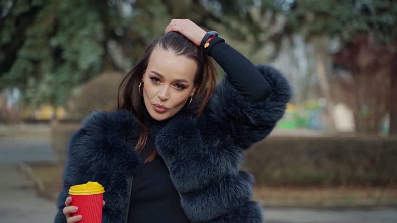 Portrait of a fashionable girl outdoors. Pretty-looking brunette model posing on camera in fur coat with a cup of coffee in the park. Slow motion.