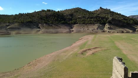 large partially dry lake with stone structure ruins in the foreground, with arid and rocky and patches of green vegetation, and a mountain in the background