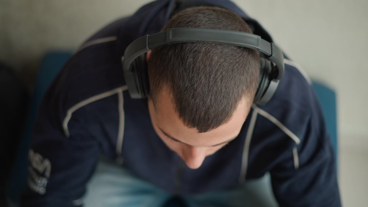 Close up of man wearing headphones and looking down with his head bent forward. subject is dressed in dark clothing seated on blue furniture against indoor background. Soft lighting creates