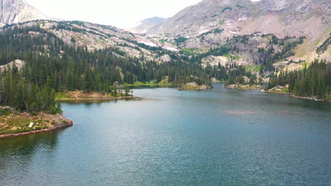 aerial drone lowering motion reveals woman standing on the end of a fallen tree log in a clear blue water lake surrounded by mountain views and a pine tree forest in nederland colorado rocky mountains