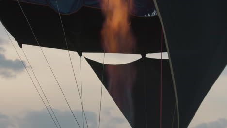 close up of hot air balloon burner releasing intense flame upward as colorful canopy begins to inflate against dramatic evening clouds, signaling critical moment in launch preparation process