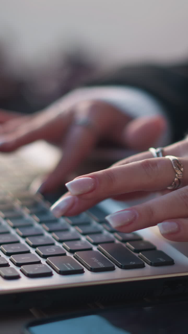 primer plano de manos con elegantes uñas cuidadas escribiendo en el teclado de la computadora portátil al aire libre, usando anillos y una cómoda manga de suéter, con una flor borrosa cerca, el reflejo visible en el teléfono colocado en la mesa