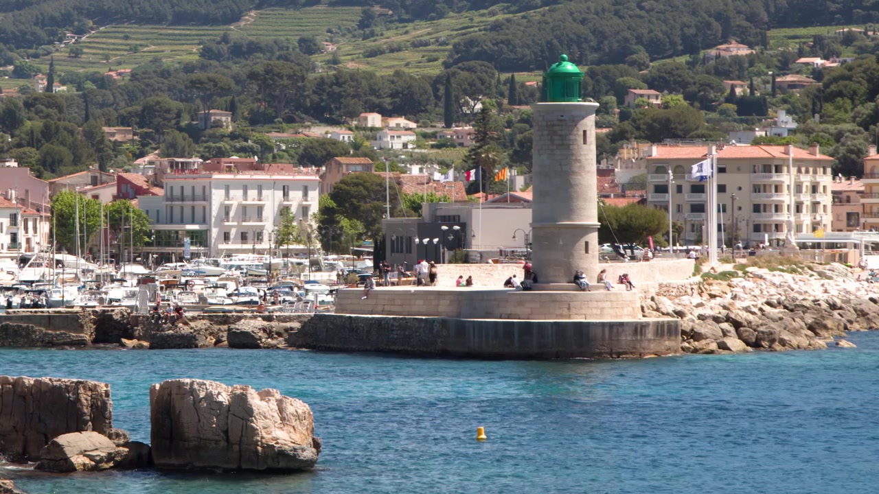 Ampia inquadratura statica di un faro in pietra che domina un porto trafficato e un villaggio costiero