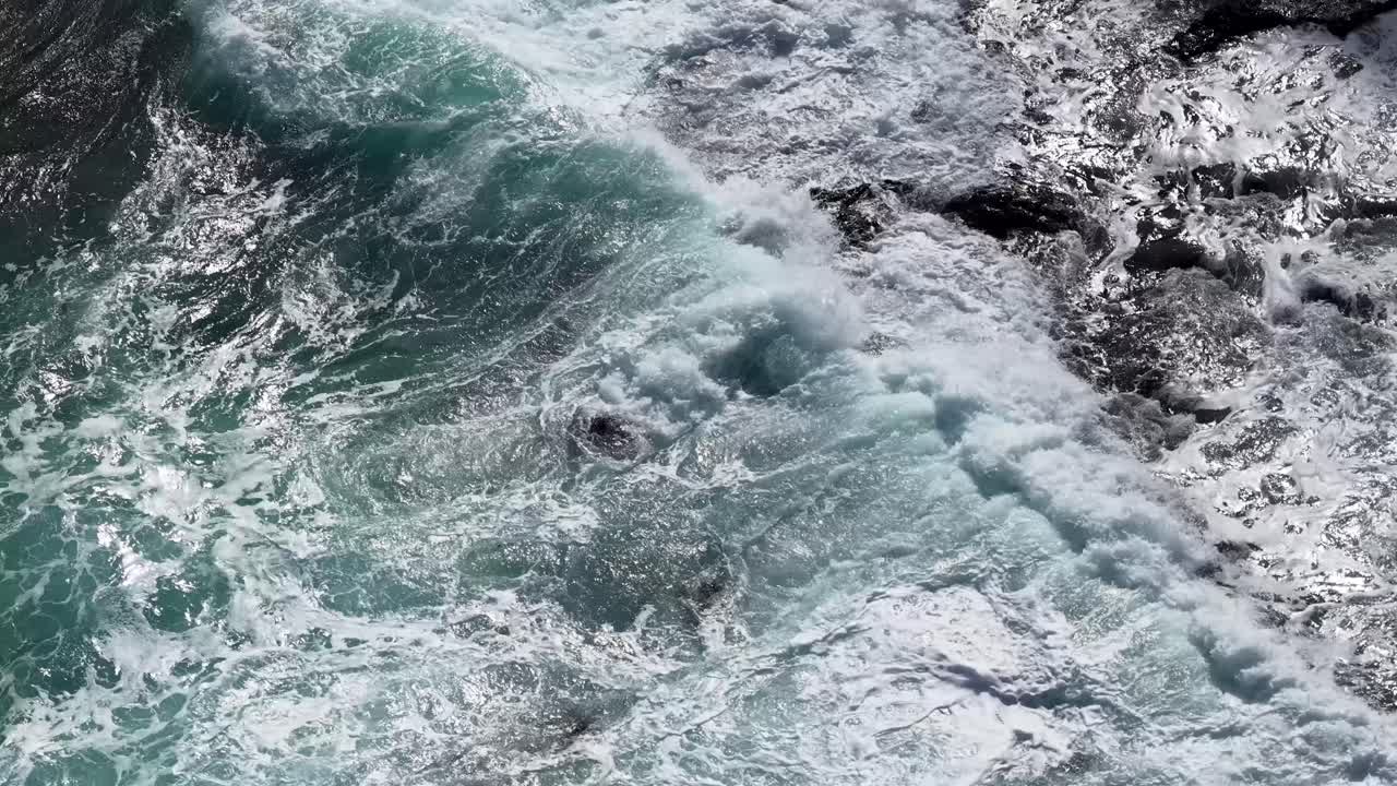Dynamic aerial view of ocean waves crashing against dark rocks, natural daylight, smooth camera movement