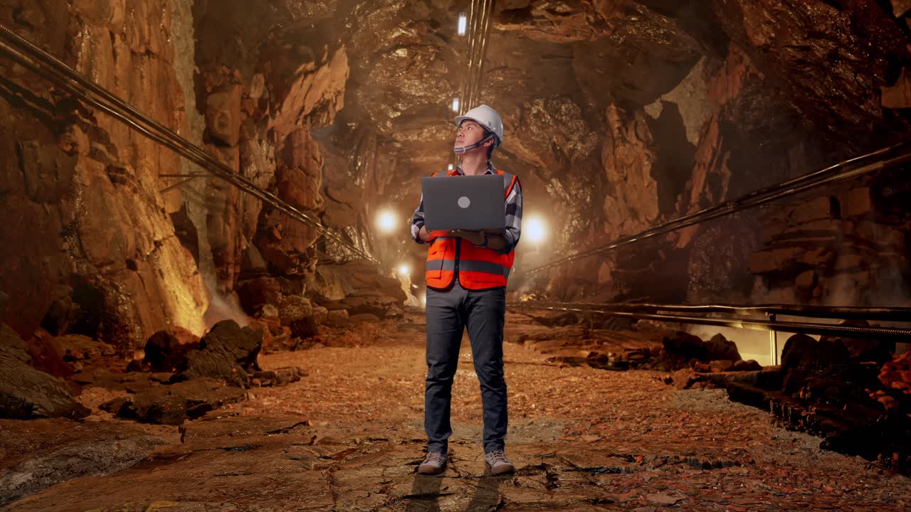 Full Body Of Asian Male Engineer With Safety Helmet Working On A Laptop And Looking Around While Standing In Underground Mine Tunnel