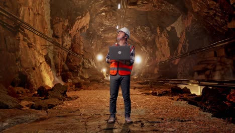 Full Body Of Asian Male Engineer With Safety Helmet Working On A Laptop And Looking Around While Standing In Underground Mine Tunnel