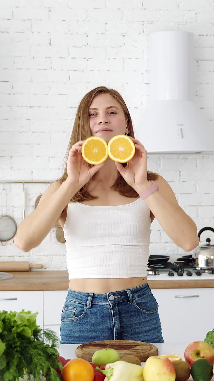 A young cheerful girl in the kitchen with fruits and vegetables is played with orange slices. The girl covers her eyes with slices of orange. Vertical video