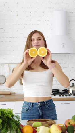 a young cheerful girl in the kitchen with fruits and vegetables is played with orange slices. the girl covers her eyes with slices of orange. vertical video