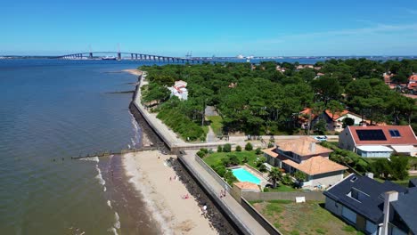 aerial drone view of a sunny coastal town featuring a sandy beach, residential homes with a swimming pool, and a large bridge spanning an estuary.
