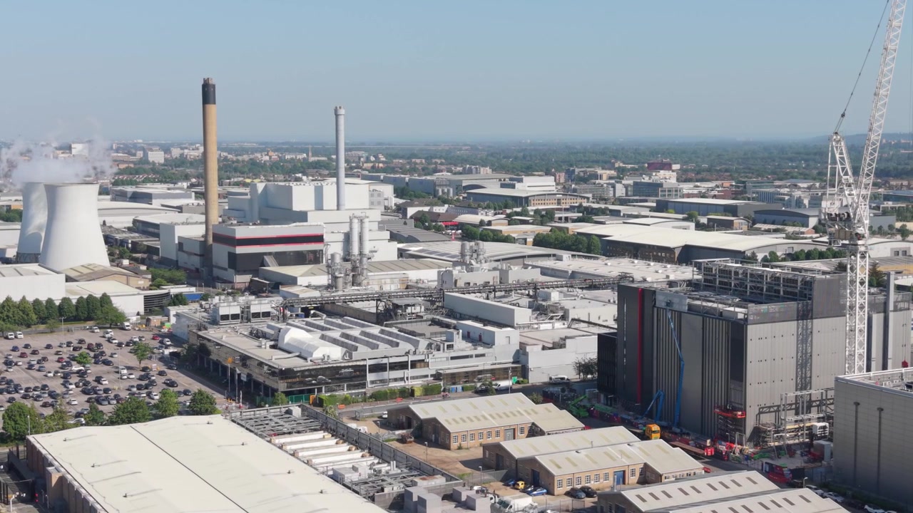 Aerial perspective reveals a vast data center complex and power facility. Cooling towers release steam while numerous buildings and full parking lots operate on a bright, sunny day.