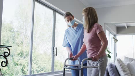 Female-health-worker-assisting-senior-woman-to-walk-with-walking-frame-at-home