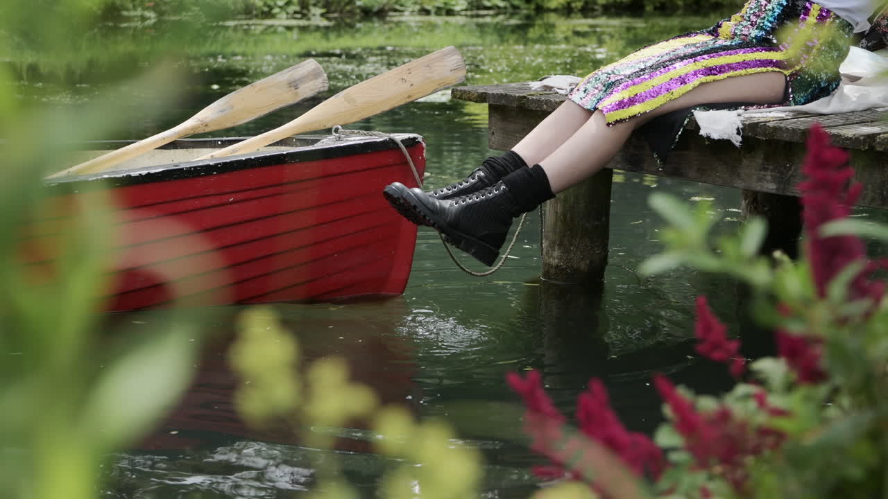 Woman relaxes on wooden jetty at small lake, dangling feet of edge, bokeh plants