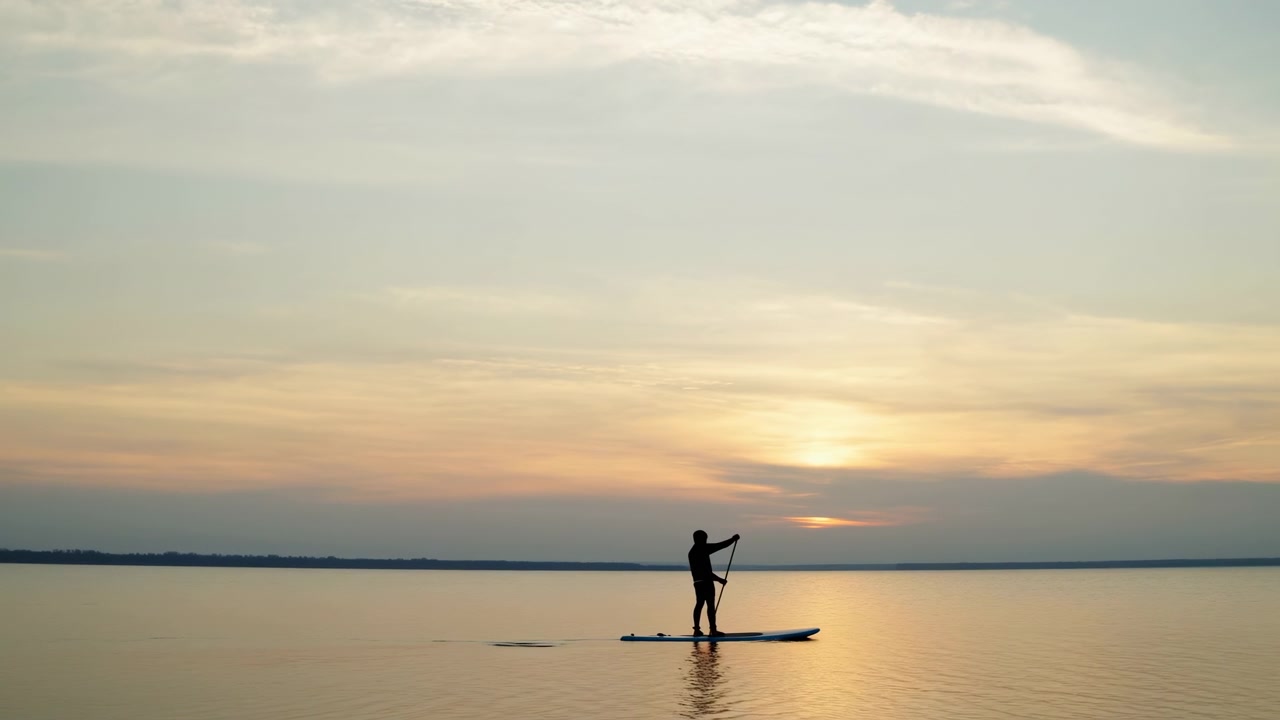 Una persona solitaria sta in piedi su una tavola da paddle al centro di un lago calmo, illuminata dal retro da un cielo al tramonto caldo con nuvole soffici e delicati riflessi sull'acqua.