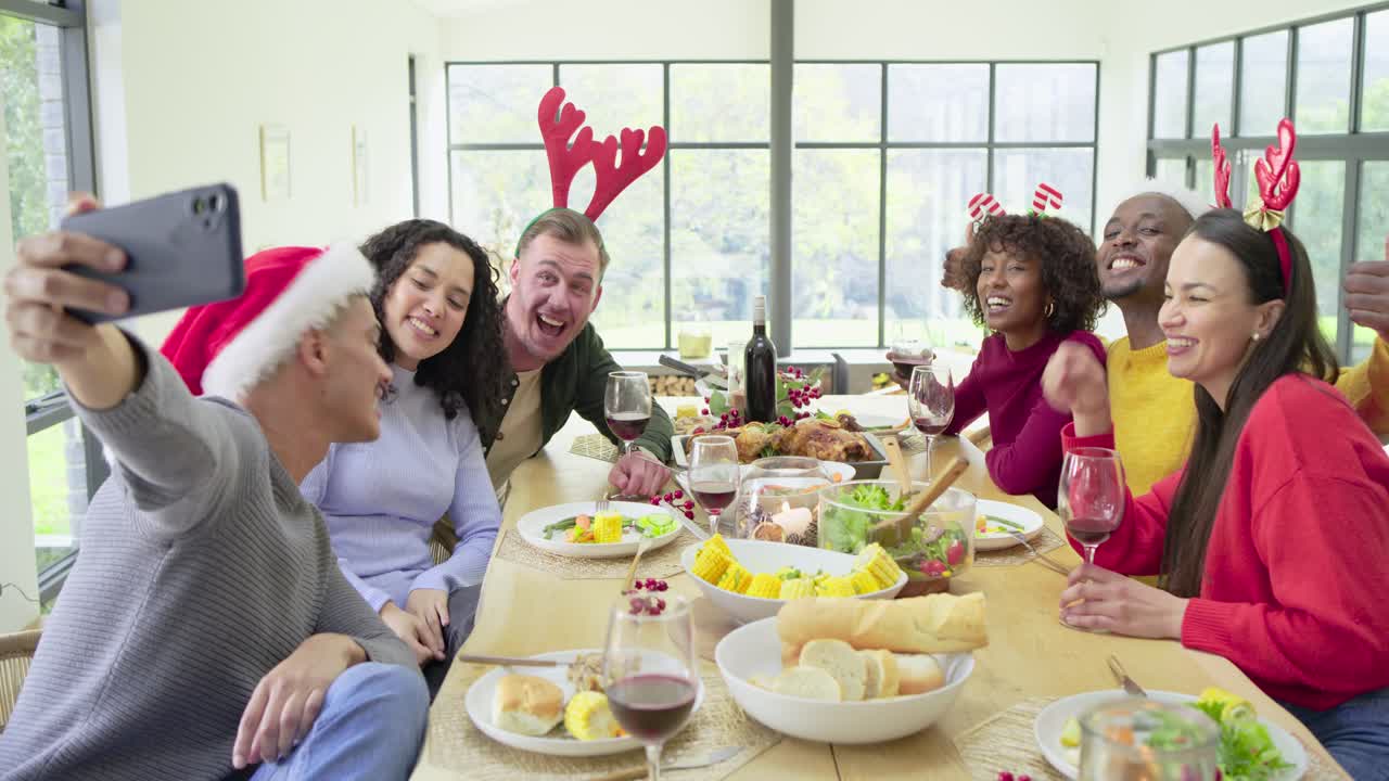 Hispanic man raising smartphone selfie at holiday dinner table with friends clinking glasses