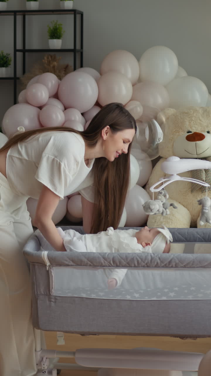 Una madre posa delicatamente il neonato nella culla accanto al letto, circondata da palloncini pastello e decorazioni per la casa.
