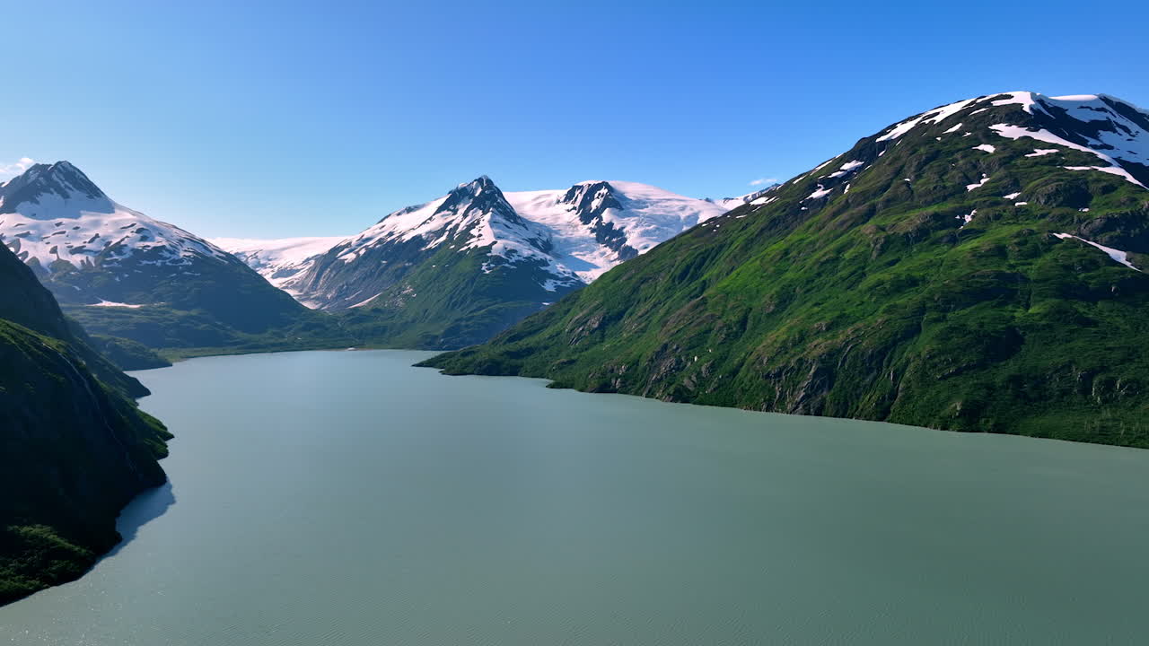Alaska summer landscape. Snow-capped peaks rise above a calm lake, surrounded by vibrant greenery under the clear blue sky of Alaska