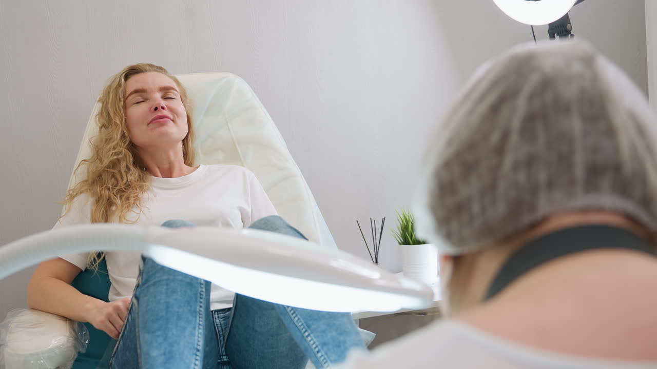 Blond client wearing white polo shirt gently smiles while reclining on chair during beauty session. Technician's back view is partially visible under bright lamp in clean, modern setting