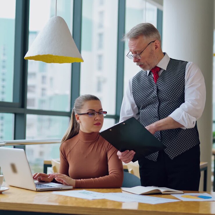 Uomo maturo e giovane donna in ufficio. Uomo d'affari che si avvicina alla segretaria seduta alla scrivania e che lavora al portatile. Concetto aziendale.