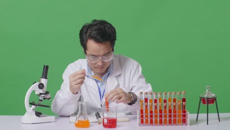 científico asiático haciendo un experimento con un tubo de ensayo y escribiendo en un cuaderno mientras trabaja en la mesa con el microscopio en el laboratorio de fondo de la pantalla verde