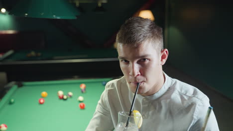 young man wearing white shirt sits on green pool table, holding cue stick and glass of lemon drink with black straw. he raises the glass and takes a sip, looking directly at the camera