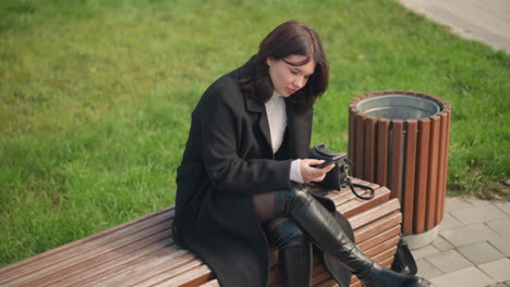 stylish young woman in black coat and white turtleneck focused on her smartphone while sitting on a park bench with crossed legs, surrounded by green grass