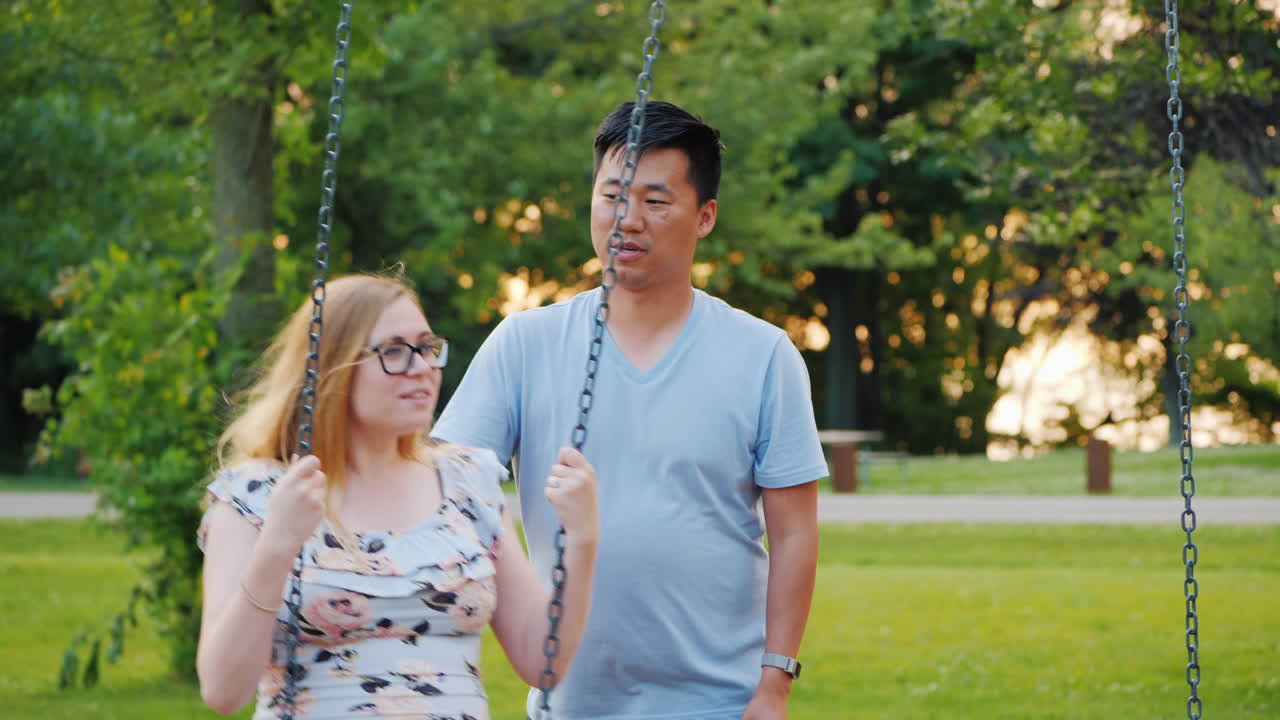 다민족 커플 - 아시아 남자와 임신한 백인 여자가 그네를 타고 행복한 젊은 가족
