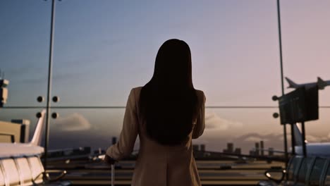 back view of asian businesswoman with rolling suitcase in boarding lounge of airline hub, talking on smartphone, waiting for flight, airport terminal with airplane takes off outside the window