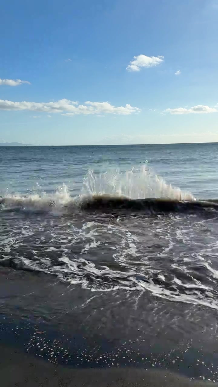 Onde oceaniche calme che si infrangono su una spiaggia di sabbia scura sotto un cielo blu