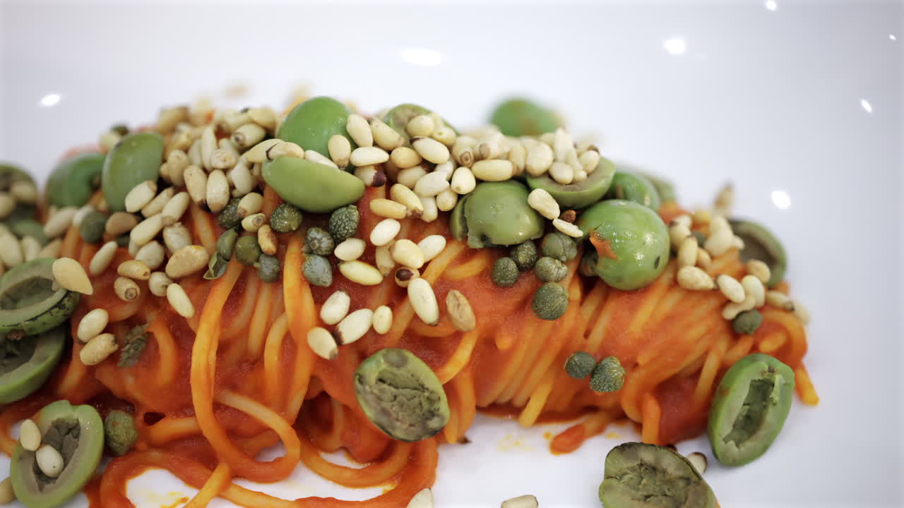Close up of a pasta dish with tomato sauce topped with green olives, seeds, and capers, presented in a modern gourmet style
