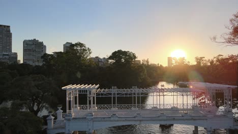 Aerial-dolly-out-of-boats-sailing-under-white-bridge-in-Rosedal-gardens-pond-at-sunset,-Palermo,-Buenos-Aires