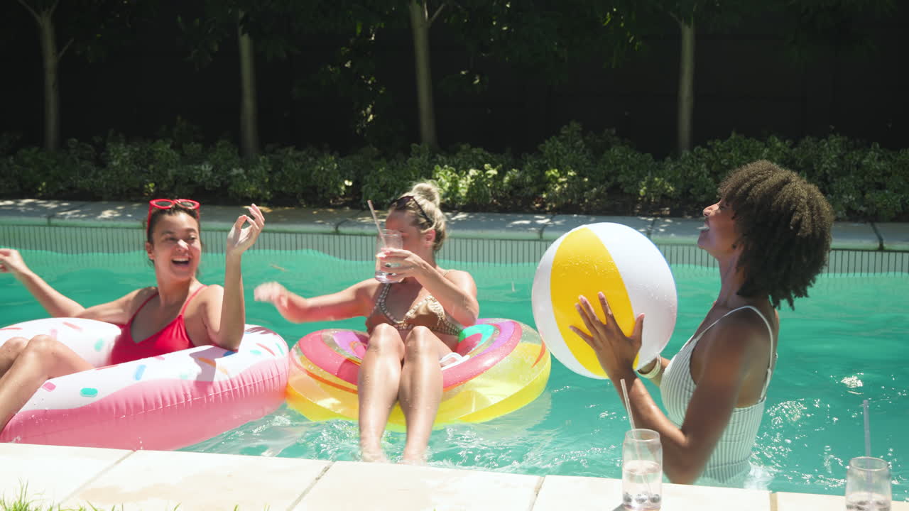 Diverse female friends enjoying pool day, floating on inflatables, playing with beach ball
