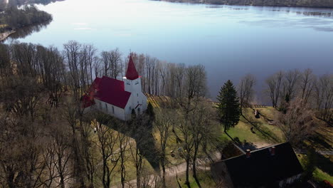 vista aérea de la iglesia luterana de lielvarde en la orilla del río daugava, iglesia blanca con techo rojo, árboles sin hojas, día soleado de primavera, cámara amplia en movimiento hacia adelante inclinada hacia abajo