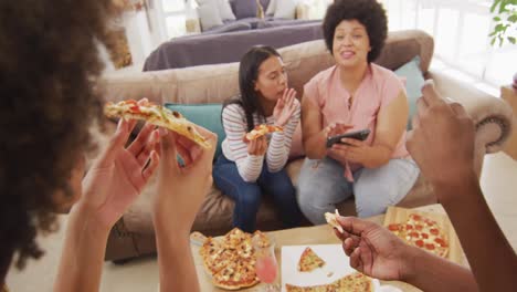 happy diverse female friends sitting on sofa in living room, eating pizza