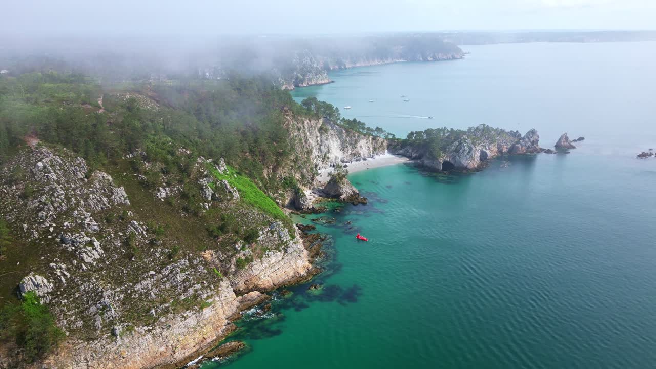 Drone shot rising over Virgin Island Beach in Crozon, Brittany, showing cliffs, rocks, turquoise sea, and mist above - France