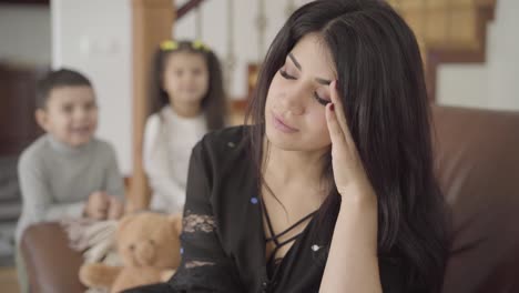 retrato de una hermosa madre de oriente medio con migraña sentada en el sillón y sus dos hijos pequeños saltando en el fondo
