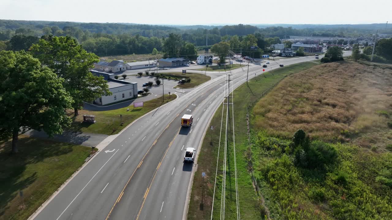 Emergency vehicle on Main Street of American suburb. Aerial tracking shot. Sunset time in small town. Strip malls and open grassy areas in USA. Summer season in Virginia