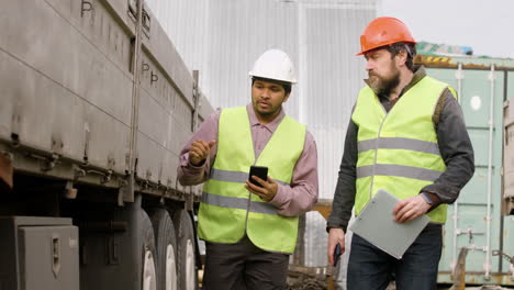 boss and worker wearing vests and safety helmets organizing a truck fleet in a logistics park while they consulting a document and walking 1