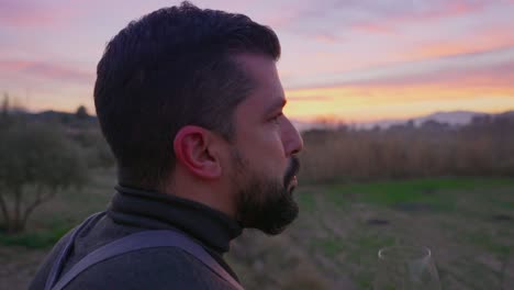 Un-Hombre-Barbudo-Bebe-Y-Prueba-Una-Copa-De-Vino-Blanco-Al-Atardecer-Bajo-Un-Cielo-Primaveral-De-Naranjas-Y-Rosas,-Con-Un-Prado-Verde