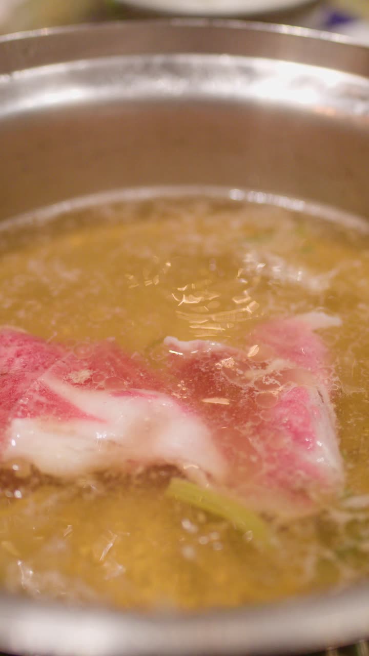 Le bacchette immergono fette di manzo marmorizzato in un brodo bollente per hotpot sotto un'illuminazione calda e interna.