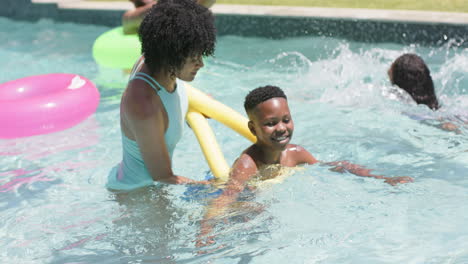 Feliz-Madre-Afroamericana-Enseñando-A-Su-Hijo-A-Nadar-Con-Fideos-Flotando-En-Una-Piscina-Soleada,-Cámara-Lenta