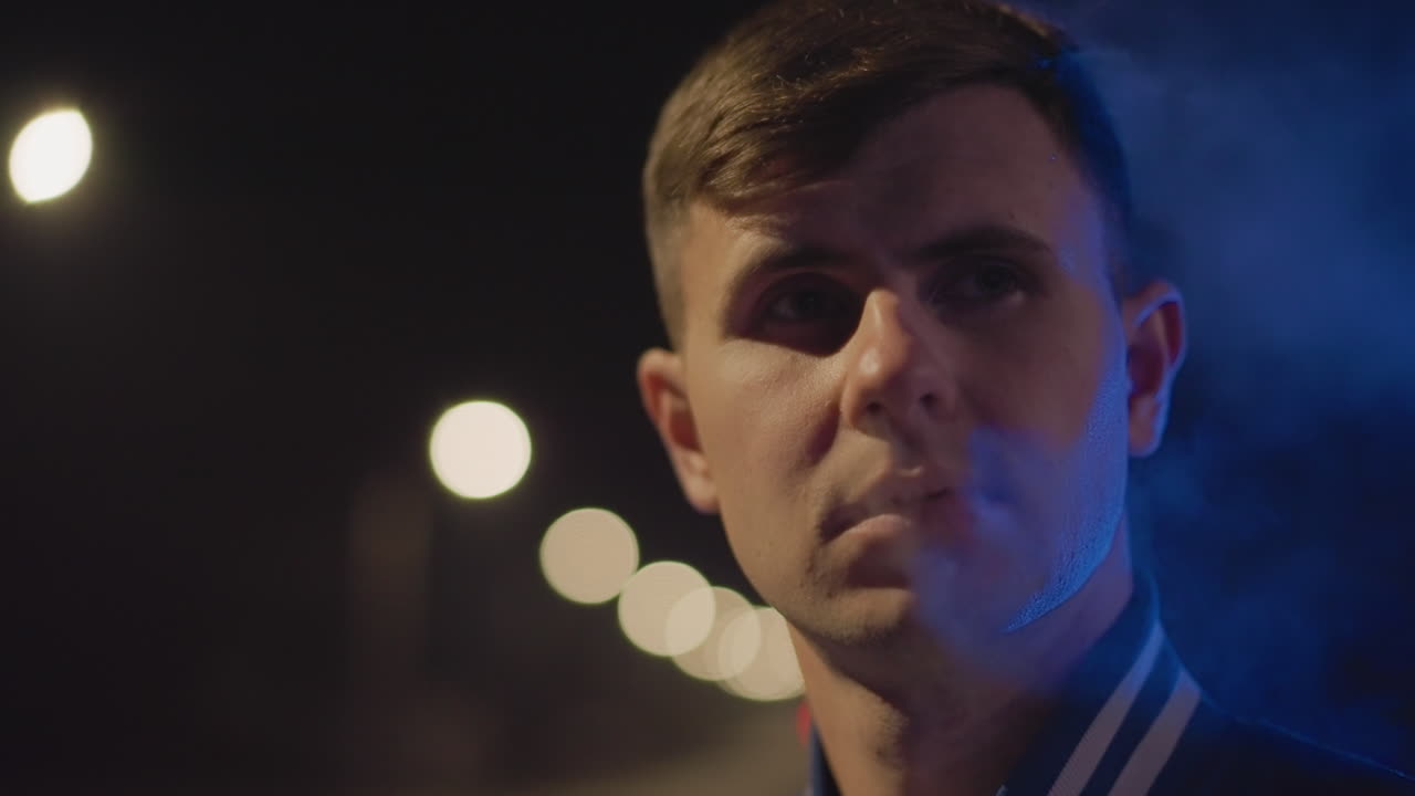 보케 아래에서 베이핑하는 생각에 잠긴 백인 남성, 도시 거리의 친밀한 야간 인물 사진, 푸른색 림 라이팅과 따뜻한 램프 불빛, 얕은 피사계 심도, 조용한 내뱉음, 사색적인 시선, 영화 같은 분위기