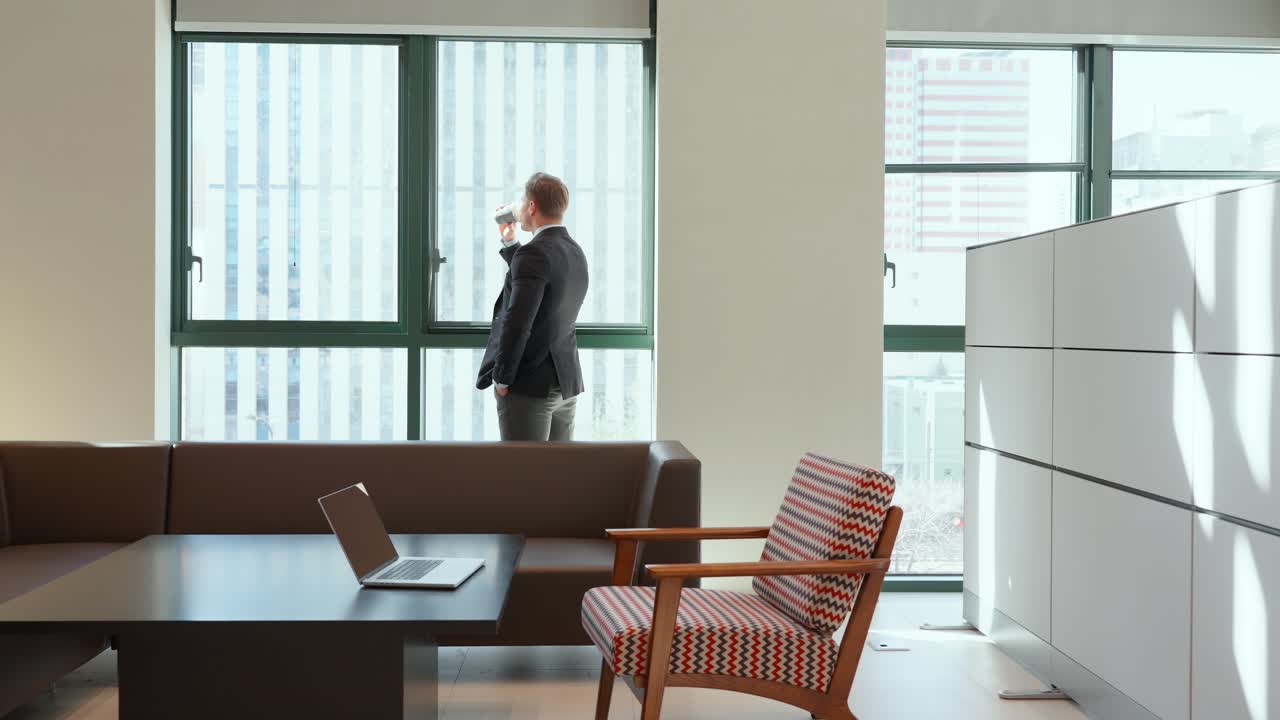 Vista posteriore di uomini d'affari che bevono caffè mattutino in piedi in modo contemplativo accanto a finestre a tutta altezza in un moderno spazio ufficio - ampia inquadratura statica.
